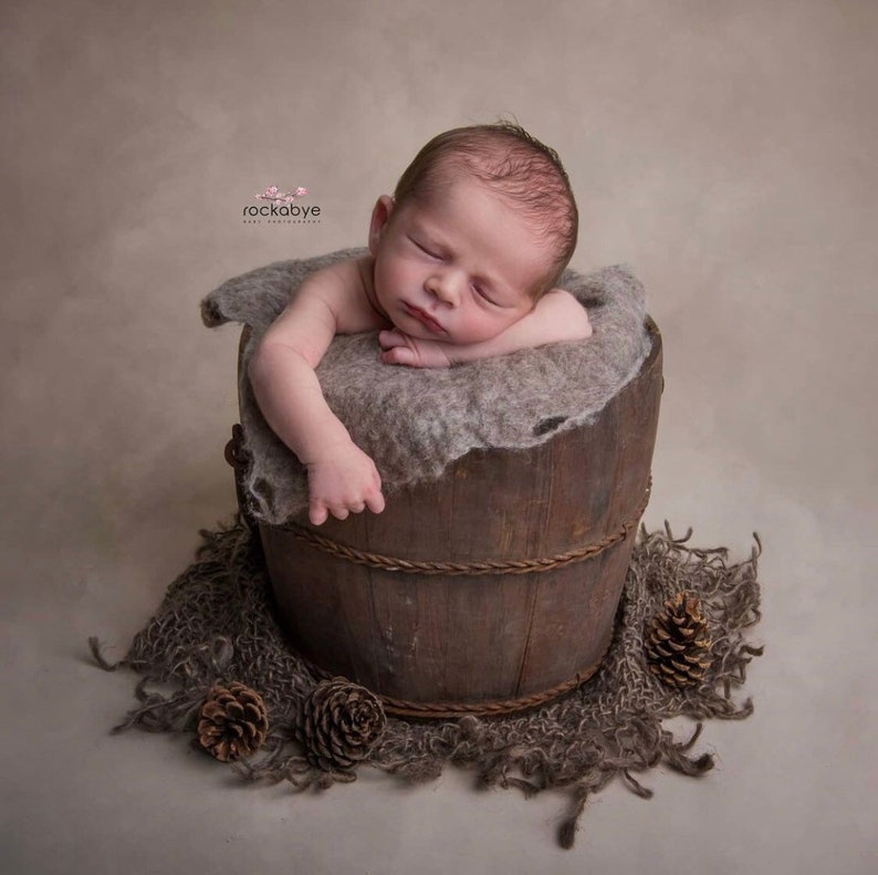 May include: A newborn baby sleeping soundly in a wooden bucket lined with a soft, grey blanket. The bucket is surrounded by a brown, textured fabric and pine cones.