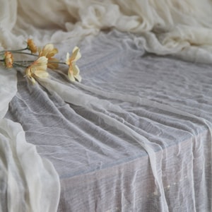 Op de afbeelding: Een wit, doorschijnend tafelkleed met een paar gele bloemen erop. De stof is over een tafel gelegd en heeft een zachte, delicate textuur.