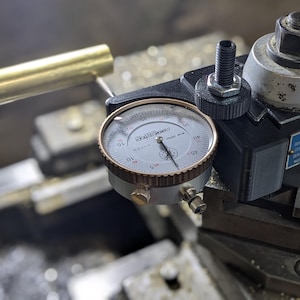 May include: A close-up of a dial indicator attached to a metal lathe. The dial indicator has a black face with white markings and a red needle. The lathe is made of metal and has a black finish.