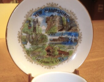May include: A white ceramic teacup and saucer set. The saucer features a colorful illustration of New Hampshire landmarks, including the text "NEW HAMPSHIRE" and "THE GRANITE STATE". The cup has a matching design.