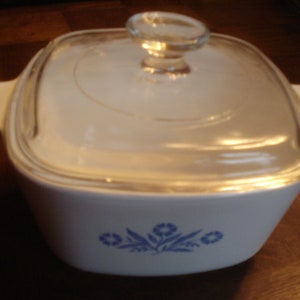 May include: White ceramic casserole dish with a glass lid and a blue floral design. The dish has a square shape with rounded corners and two handles on the sides.