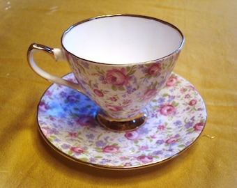 May include: A floral teacup and saucer set with a white background and pink and purple flowers. The cup and saucer have a gold trim.