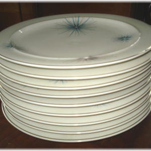 May include: A stack of ten white ceramic dinner plates with a gold rim and a blue and white starburst design.