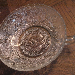 May include: A clear glass bowl with a decorative pattern and a single handle. The bowl has a raised center section with a star-shaped design.