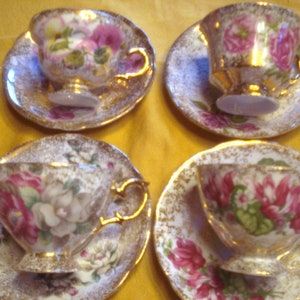 May include: Four vintage teacups and saucers with floral patterns and gold accents. The cups and saucers have a white background with gold flecks and a floral design in shades of pink, red, and green.