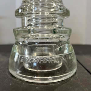May include: A clear glass insulator with a rounded top and a wide base. The base has the text "Hemingray Glass" embossed on it.