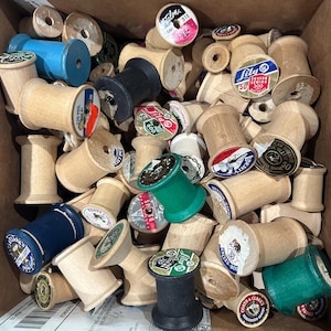 May include: A box filled with wooden spools of thread. The spools are various sizes and have different colored thread. Some of the spools have labels with brand names and thread information, such as "Talon", "Lily", "Liberty", and "Coats & Clarkes".