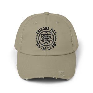 May include: A distressed beige baseball cap with a black embroidered logo that reads "Arizona Bay Swim Club" and features a stylized star with a spiral inside.