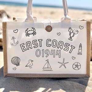 May include: A white tote bag with a jute border and cream-colored handles. The bag features black line drawings of nautical elements and the text "EAST COAST 01944". A wooden button secures the top.