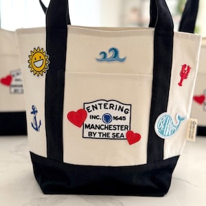 Puede incluir: Bolso tote color crema con asas y base negras. El bolso presenta parches bordados de un sol sonriente, un ancla, una ola, una langosta, una ballena y corazones. El texto "Entering Manchester by the Sea" también está bordado.