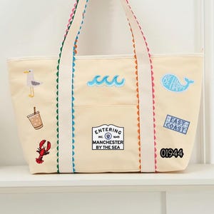 Pode incluir: Bolsa tote creme com acabamentos e alças coloridas. A bolsa apresenta patches bordados de uma gaivota, ondas, uma baleia, uma lagosta, uma chávena de café e o texto "Entering Manchester By The Sea" e "East Coast".