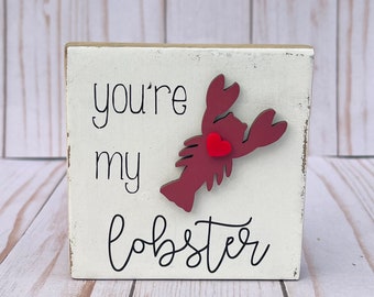 May include: White wooden sign with the text "you're my lobster" in black cursive font. A red lobster with a red heart is attached to the sign.