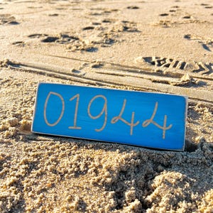 May include: A blue wooden sign with the number 01944 written in white paint. The sign is lying on a sandy beach.