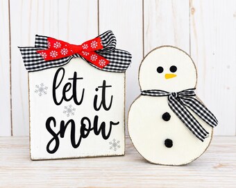 Könnte beinhalten: Ein rechteckiges weißes Holzschild mit dem schwarzen Schriftzug let it snow und silbernen Schneeflocken, verziert mit einer rot-schwarz karierten Schleife, neben einer weißen Schneemannfigur aus Holz mit schwarz-weiß kariertem Schal.