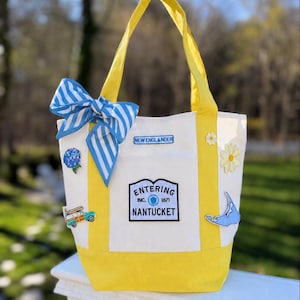 Puede incluir: Un bolso de lona blanco con asas y base amarillas, decorado con un lazo de rayas azules y blancas. Los parches incluyen una hortensia azul, un coche familiar, una etiqueta New Englander, un letrero de Entering Nantucket, una margarita y un mapa.