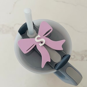 May include: A gray travel mug with a pink bow and the letter "S" on it. The mug has a lid with a straw hole and a handle.