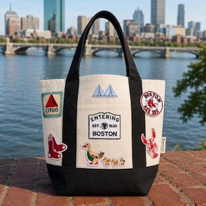 Puede incluir: Un bolso de tela color crema con asas y base negras. El bolso presenta parches bordados que incluyen un letrero de Citgo, un puente, un logotipo de los Boston Red Sox, un calcetín rojo, una langosta, un pato con patitos y un parche que dice Entering Est. 1630 Boston.