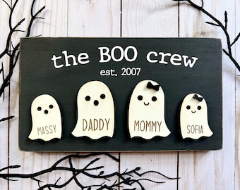 Puede incluir: Un letrero rectangular de madera gris oscuro con el texto blanco "the BOO crew" y "est. 2007" sobre cuatro siluetas de fantasmas de madera clara. Los fantasmas tienen ojos y bocas negros, y los nombres "MASSY", "DADDY", "MOMMY" y "SOFIA" impresos.