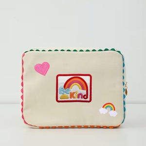 Pode incluir: Bolsa retangular creme com um design de arco-íris e coração. A bolsa apresenta uma borda ondulada vermelha, rosa, verde e azul. A frente da bolsa tem um coração rosa e um arco-íris com as palavras "Be Kind".