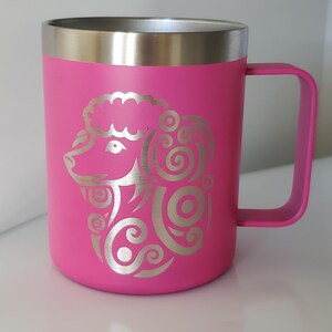 Könnte beinhalten: Ein leuchtend pinkfarbener Reisebecher mit silbernem Rand und Griff. Der Becher zeigt ein silbernes Zierdesign eines Widderkopfes. Der Becher ist für ca. 400 ml Flüssigkeit ausgelegt.