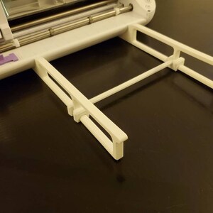 K&ouml;nnte beinhalten: Eine wei&szlig;e Materialst&uuml;tzverl&auml;ngerung aus Kunststoff, die an einer wei&szlig;en elektronischen Schneidemaschine befestigt ist. Das Tablett verf&uuml;gt &uuml;ber zwei parallele, rechteckige Seitenarme, die durch eine horizontale Querstange verbunden sind, um Materialien flach zu halten.