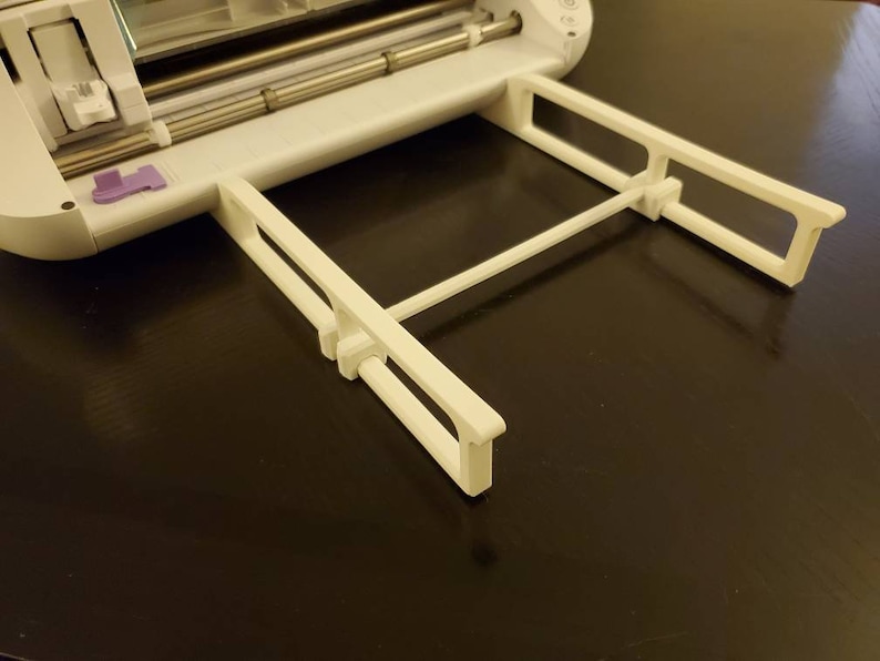 K&ouml;nnte beinhalten: Eine wei&szlig;e Materialst&uuml;tzverl&auml;ngerung aus Kunststoff, die an einer wei&szlig;en elektronischen Schneidemaschine befestigt ist. Das Tablett verf&uuml;gt &uuml;ber zwei parallele, rechteckige Seitenarme, die durch eine horizontale Querstange verbunden sind, um Materialien flach zu halten.