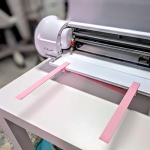 May include: A white Juliet electronic cutting machine featuring a horizontal feed slot and a metallic internal carriage. Two light pink plastic support arms are attached to the front of the machine, extending outward to provide a flat surface for material.