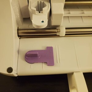 K&ouml;nnte beinhalten: Eine wei&szlig;e elektronische Schneidemaschine mit einer horizontalen Metallstange und einem wei&szlig;en Kunststoff-Werkzeughalter. Ein flaches, violettes, T-f&ouml;rmiges Kunststoffteil mit einer erh&ouml;hten rechteckigen Lasche liegt auf der Basis.