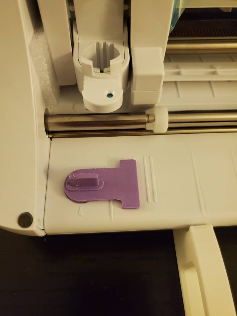 K&ouml;nnte beinhalten: Eine wei&szlig;e elektronische Schneidemaschine mit einer horizontalen Metallstange und einem wei&szlig;en Kunststoff-Werkzeughalter. Ein flaches, violettes, T-f&ouml;rmiges Kunststoffteil mit einer erh&ouml;hten rechteckigen Lasche liegt auf der Basis.