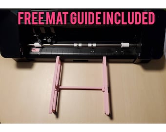 May include: A black electronic cutting machine with a metallic roller bar and white rollers, featuring a light pink plastic mat guide attachment positioned in front. The text FREE MAT GUIDE INCLUDED is displayed in bold pink letters at the top.