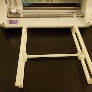 K&ouml;nnte beinhalten: Eine wei&szlig;e elektronische Schneidemaschine mit einer metallischen Rollenstange und einem violetten Riegel, ausgestattet mit einer wei&szlig;en Kunststoff-Ablageverl&auml;ngerung an der Vorderseite. Das Ger&auml;t verf&uuml;gt &uuml;ber ein Bedienfeld mit verschiedenen Tasten.