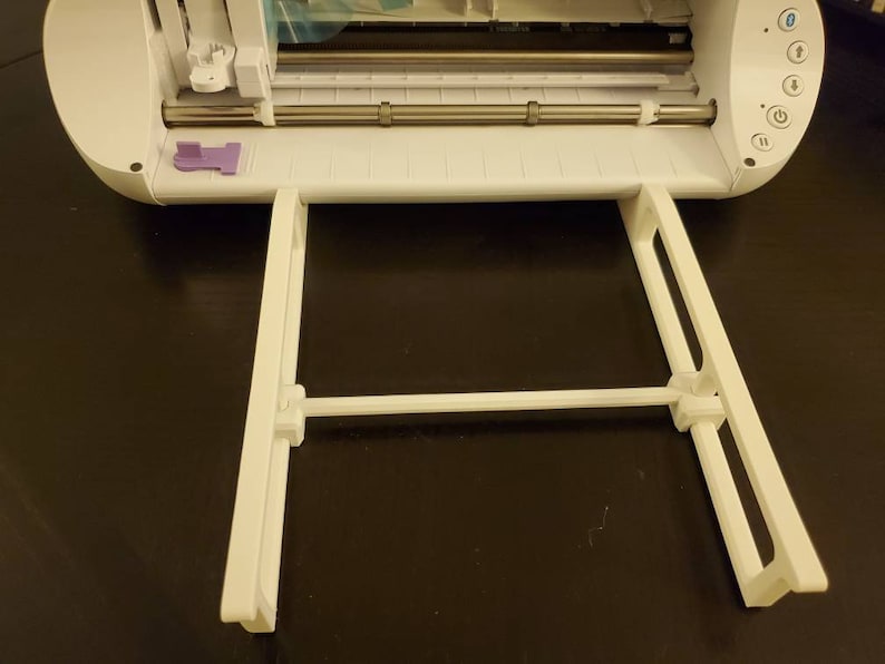 K&ouml;nnte beinhalten: Eine wei&szlig;e elektronische Schneidemaschine mit einer metallischen Rollenstange und einem violetten Riegel, ausgestattet mit einer wei&szlig;en Kunststoff-Ablageverl&auml;ngerung an der Vorderseite. Das Ger&auml;t verf&uuml;gt &uuml;ber ein Bedienfeld mit verschiedenen Tasten.