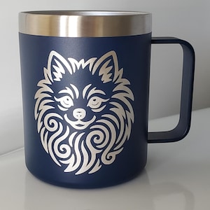 Könnte beinhalten: Marineblauer Reisebecher mit silbernem Rand und Griff. Der Becher zeigt eine detaillierte, cremefarbene Illustration eines Hundegesichts. Das Hundegesicht ist auf dem Becher zentriert, mit einem lächelnden Ausdruck.