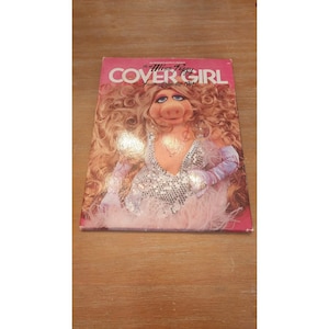 May include: A 1981 Miss Piggy Cover Girl fantasy calendar featuring a large illustration of the character with voluminous blonde hair, wearing a silver sequined top and matching gloves against a pink background with bold white text.