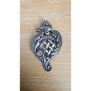 May include: A silver-toned metal brooch featuring an intricate openwork lattice design at the center, surrounded by ornate, swirling scrollwork and small spherical accents. The piece has a polished finish with darkened recessed areas for contrast.