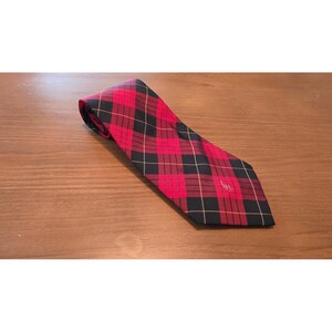 May include: A necktie featuring a red and black plaid tartan pattern with thin white intersecting lines. The fabric has a smooth texture and a pointed end. A small, embroidered logo is visible near the bottom of the tie, adding detail.