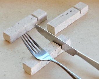 May include: A set of three white ceramic knife and fork rests. The rests are made of a rough, textured material and have a rectangular shape. A silver fork and knife are placed on the rests.