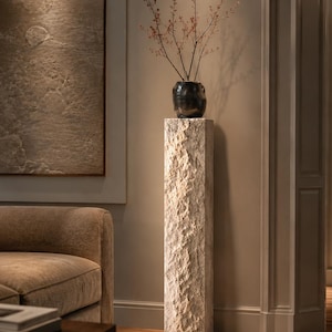 May include: A tall, rectangular pedestal with a rough, chiseled stone texture in a light beige color. Resting on top is a dark, rounded ceramic vase containing thin, bare branches with small, reddish-brown buds, creating a vertical display.