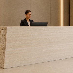 May include: A long, rectangular reception desk made of beige travertine stone. The front and top surfaces are smooth with horizontal veining, while the left side features a raw, porous, and textured natural stone edge with a jagged finish.