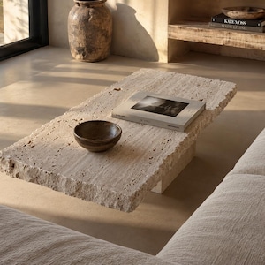 May include: A rectangular coffee table made of beige travertine stone with a rough, natural chiseled edge. The table features a flat, porous surface and a solid block base. A small dark brown bowl and a book with a light cover rest on the top.