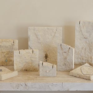 May include: A collection of jewelry display stands made of textured, light-colored stone. Various necklaces and a ring are displayed on the stands. The stands vary in size and shape, creating a visually appealing arrangement. The background is a neutral beige.