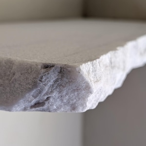 Peut inclure: Gros plan sur le bord d'une étagère en pierre blanche texturée. Le bord présente une surface rugueuse et inégale avec de petites inclusions sombres. L'étagère semble faire partie d'une structure plus grande.