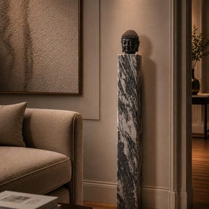 May include: A dark gray Buddha head sculpture resting atop a tall, rectangular pedestal. The pedestal features a striking black and white marbled stone pattern with vertical veining, standing upright on a wooden floor in a room setting.