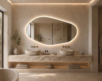 May include: An asymmetrical, organic-shaped wall mirror with integrated LED backlighting, positioned above a double vanity featuring two rough-hewn stone vessel sinks, brass wall-mounted faucets, and a textured beige stone countertop.