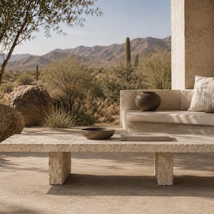 May include: A rectangular coffee table made of light-colored, textured stone with a rough-hewn edge and two thick, block-style legs. The table is positioned outdoors and holds a small, dark, round bowl and a flat, rectangular object on its surface.