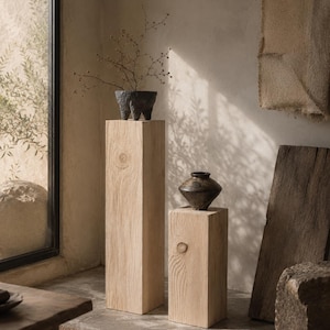 May include: Two light-colored wooden pedestals with a square profile and visible wood grain. The taller pedestal holds a dark, textured, three-legged bowl with thin branches, while the shorter pedestal supports a dark, wide-rimmed ceramic vase.