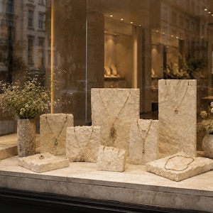 May include: A jewelry display featuring multiple gold necklaces and rings arranged on rough-hewn, light beige stone blocks. Two stone vases containing dried flowers are placed on the sides of the display, which is set behind a glass window.
