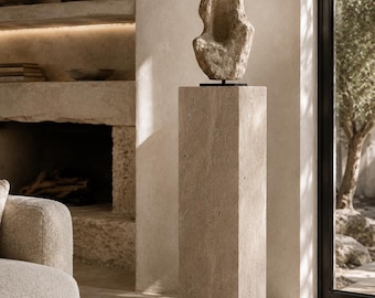 May include: A tall, rectangular beige travertine stone pedestal stand with a porous, textured surface. Resting on top of the pedestal is an abstract, organic-shaped beige stone sculpture mounted on a thin black metal base and rod.