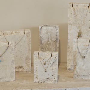 May include: A display of silver necklaces on textured, off-white stone stands. The necklaces feature various pendant designs, including a heart and a small charm. The stands are arranged on a light-colored surface, showcasing the jewelry.