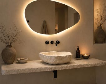 May include: An irregular, pebble-shaped wall mirror with integrated warm LED backlighting, positioned above a rough-hewn stone vessel sink and a thick, natural stone floating vanity shelf featuring matte black wall-mounted plumbing fixtures.